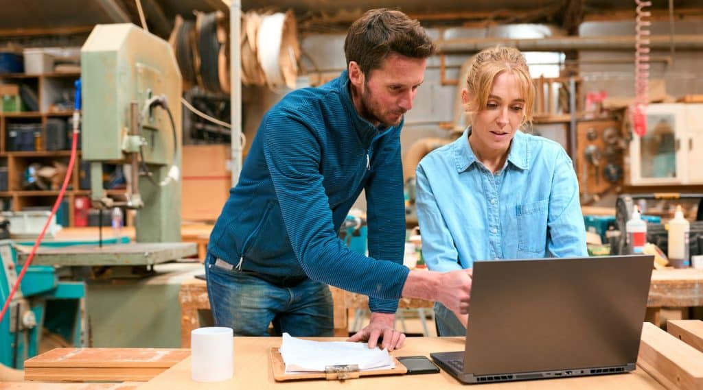Male and female business co-owner working on a laptop on a timber desk inside a carpentry workshop