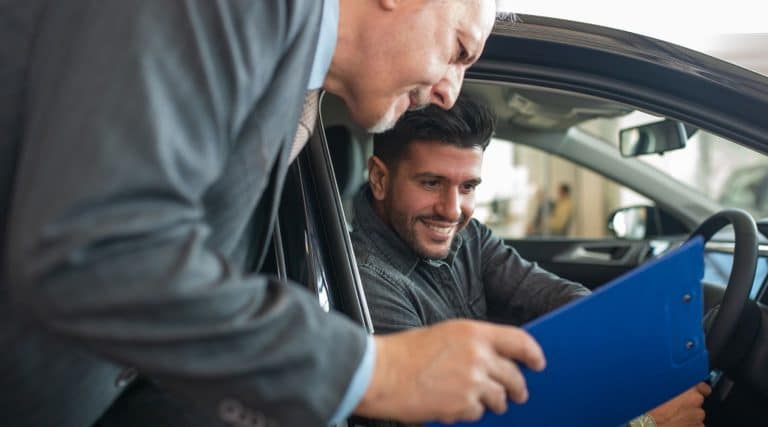 Man sitting in a car at a dealership looking at a clipboard with a salesman